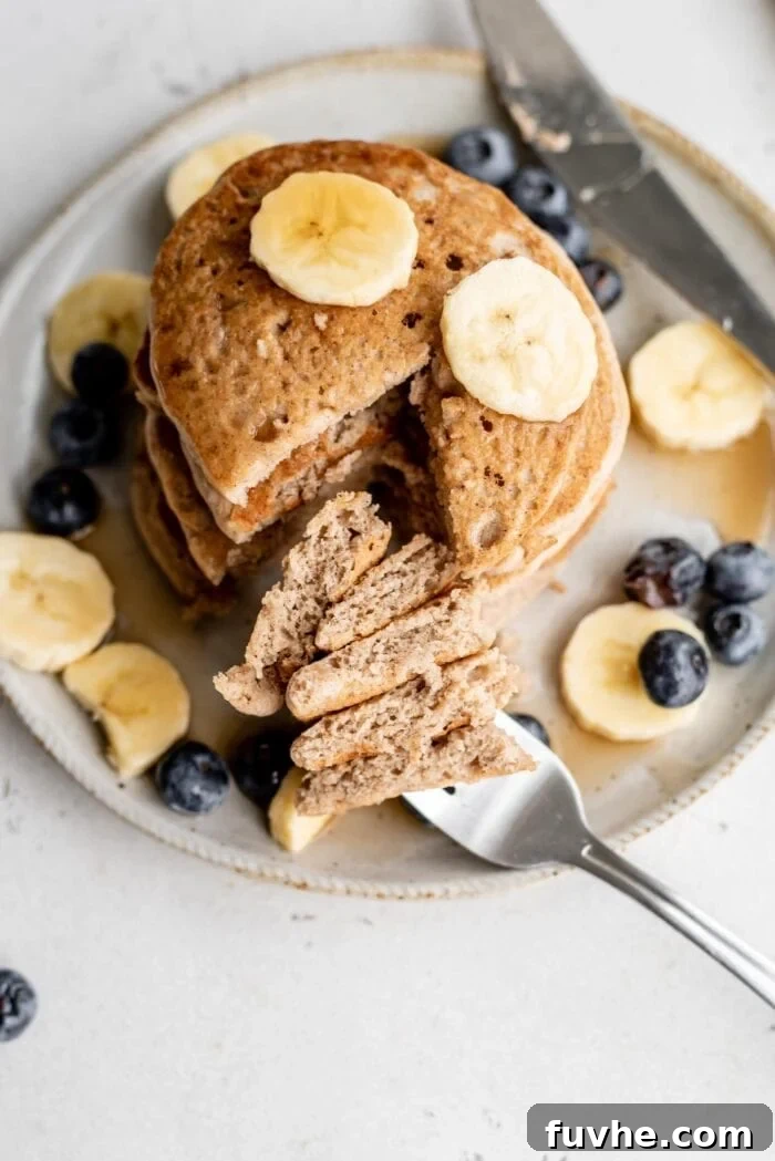 Plant-Based Buckwheat Power Pancakes 7 A beautifully presented stack of fluffy buckwheat pancakes, sliced to reveal their tender interior, gorgeously topped with vibrant fresh blueberries and golden banana slices, drizzled with syrup on a pristine white plate.