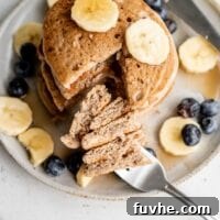 A beautifully presented stack of buckwheat pancakes, sliced to reveal their fluffy interior, topped with vibrant blueberries and golden banana slices on a white plate.