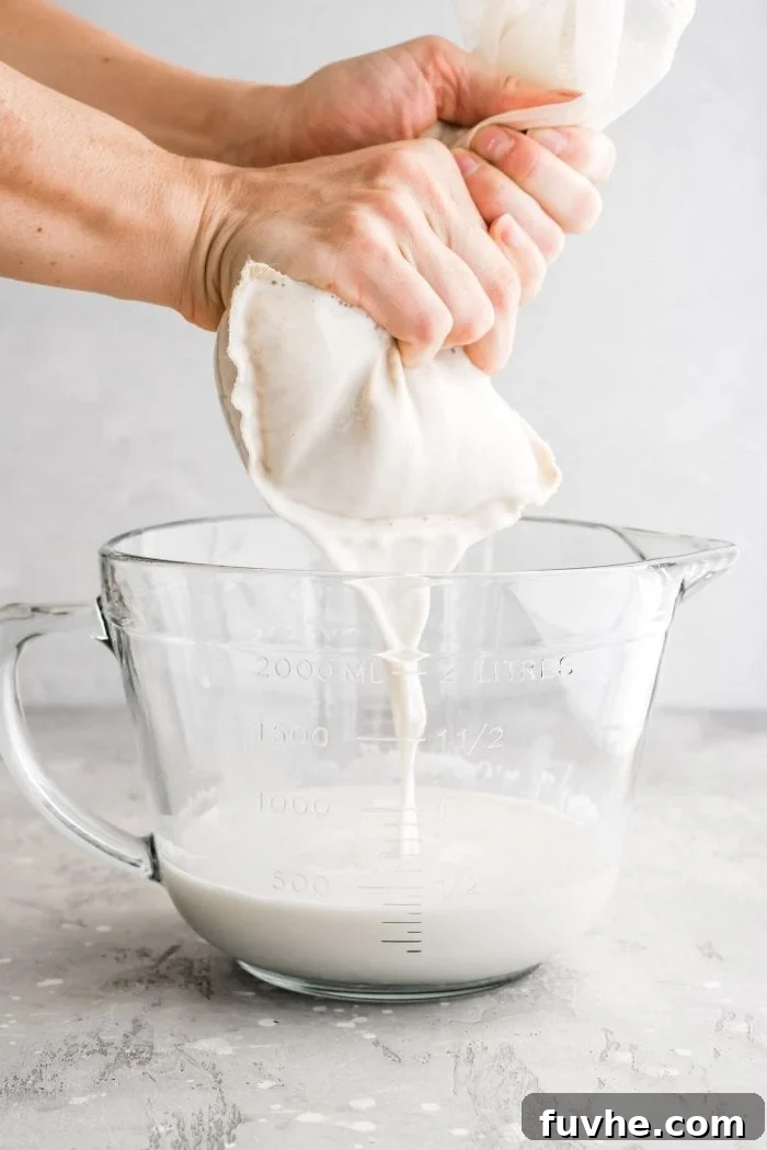 Carefully straining the blended oat milk through a nut milk bag into a glass measuring cup for a perfectly smooth consistency.