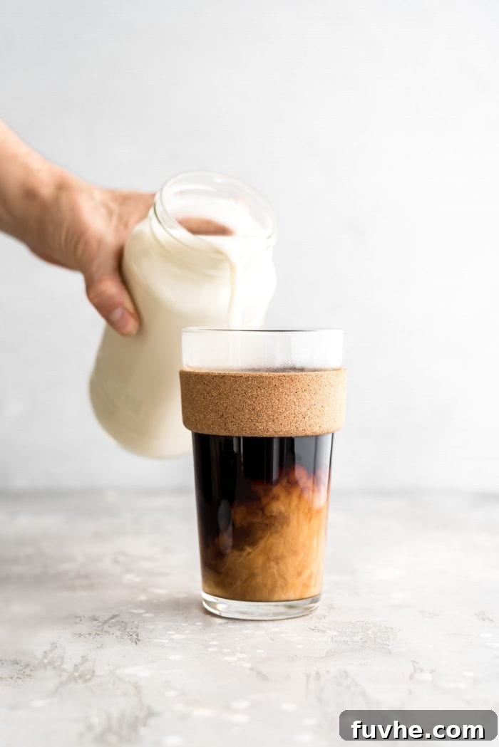 Pouring homemade oat milk into a glass of hot coffee, creating a beautiful swirl.