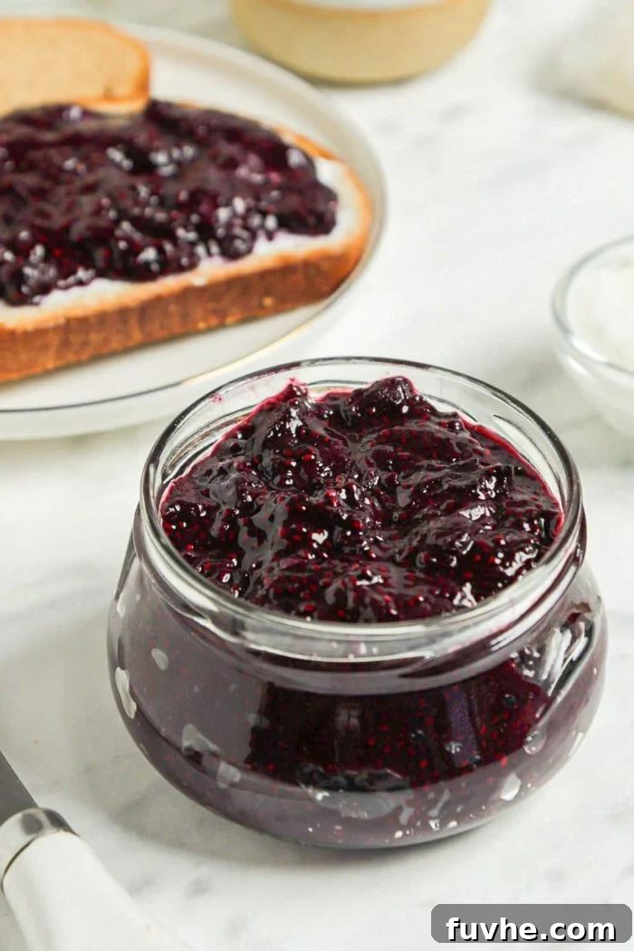 Whip Up Easy Berry Chia Seed Jam in Minutes 2 Small jar of blueberry chia seed jam with a piece of bread topped with jam in the background, showcasing its homemade appeal.