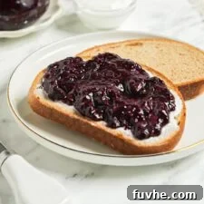 Piece of toast topped with blueberry chia jam. Jar of blueberry jam and a mug of coffee in the background.