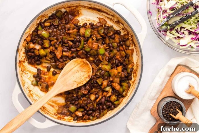 A rich BBQ black bean mixture simmering gently in a saucepan, with a wooden spoon resting in it, showing a thick and saucy consistency.