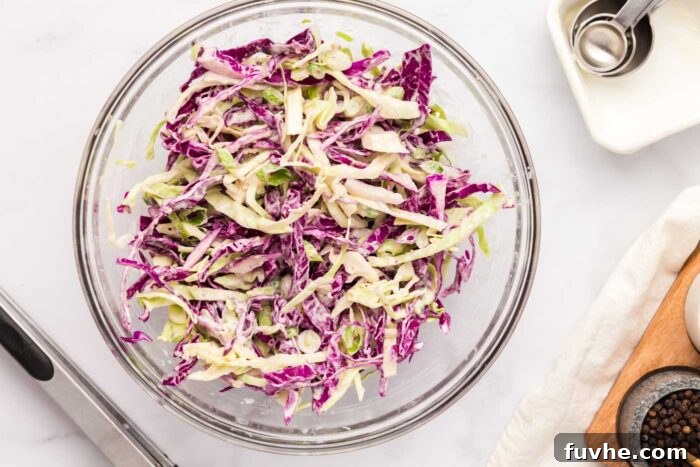 Vibrant red and green cabbage slaw, coated in a creamy dressing, being mixed in a glass bowl.