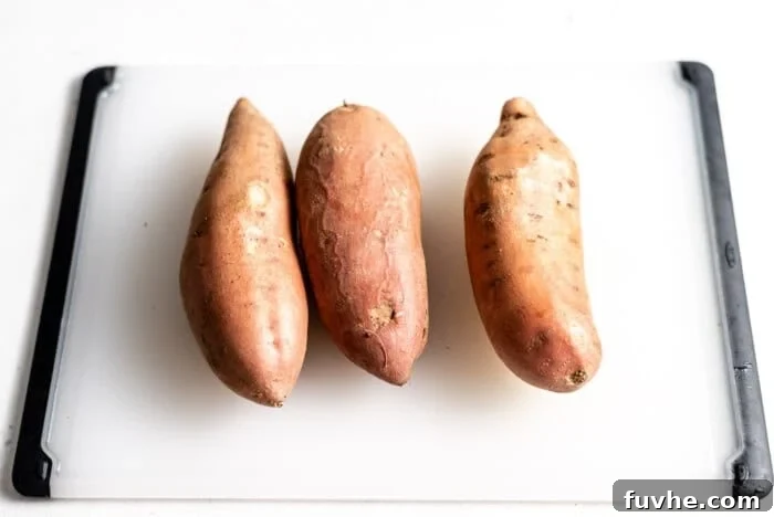 Three whole, unpeeled sweet potatoes resting on a wooden cutting board, ready for preparation.