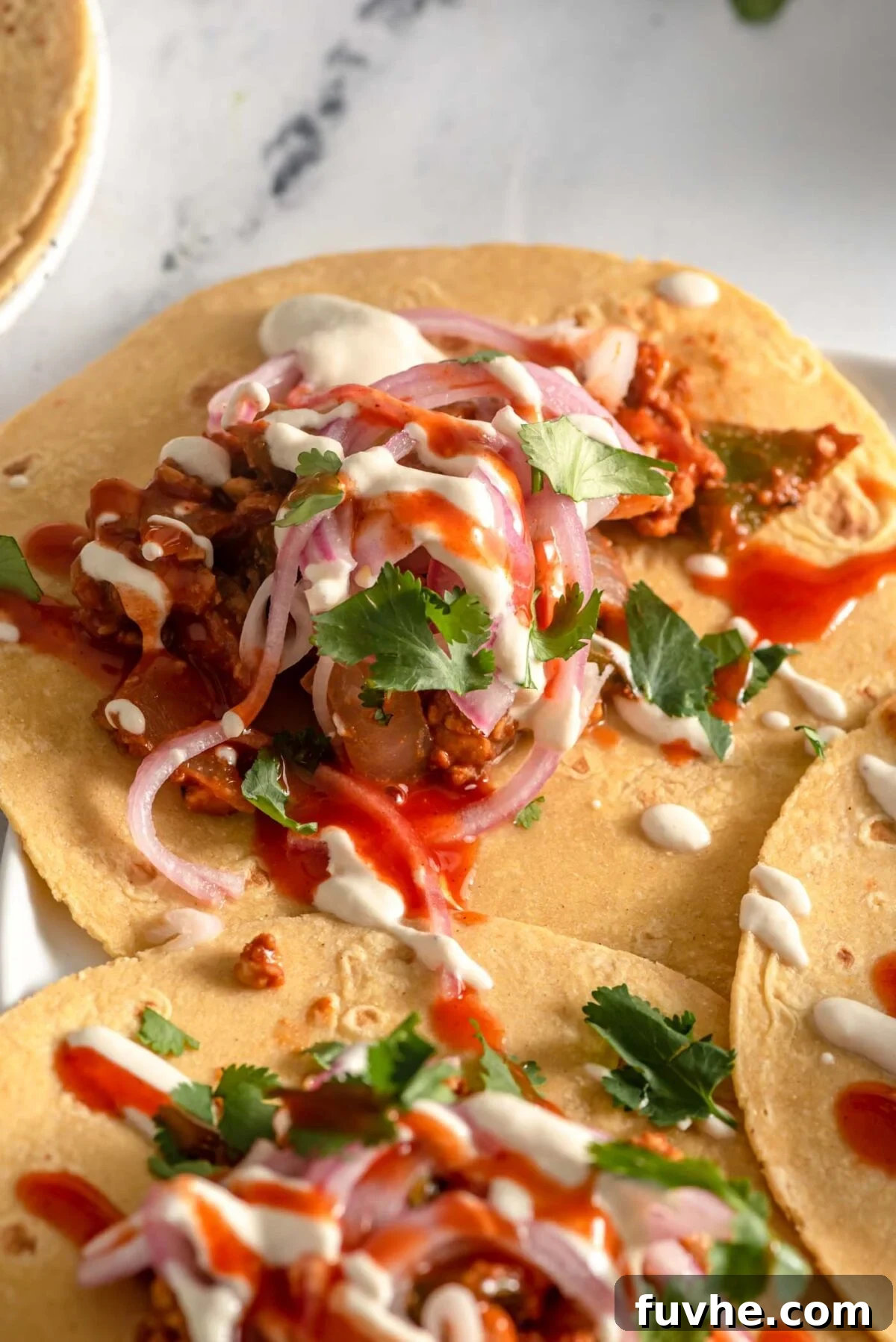 Savory Tempeh Tacos 9 Three beautifully assembled tempeh tacos adorned with vibrant pickled red onion, a generous drizzle of cashew cream sauce, and a hint of hot sauce, artfully arranged on a plate.