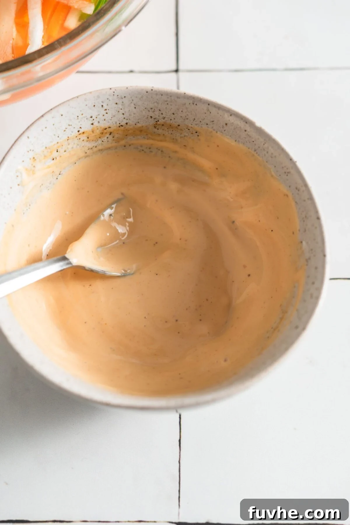 A small bowl filled with homemade spicy mayonnaise, accompanied by a spoon, on a rustic surface.