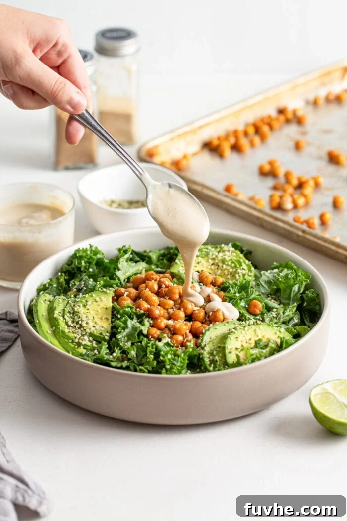 A hand drizzling a spoonful of creamy tahini dressing over a vibrant chickpea avocado kale salad in a bowl.