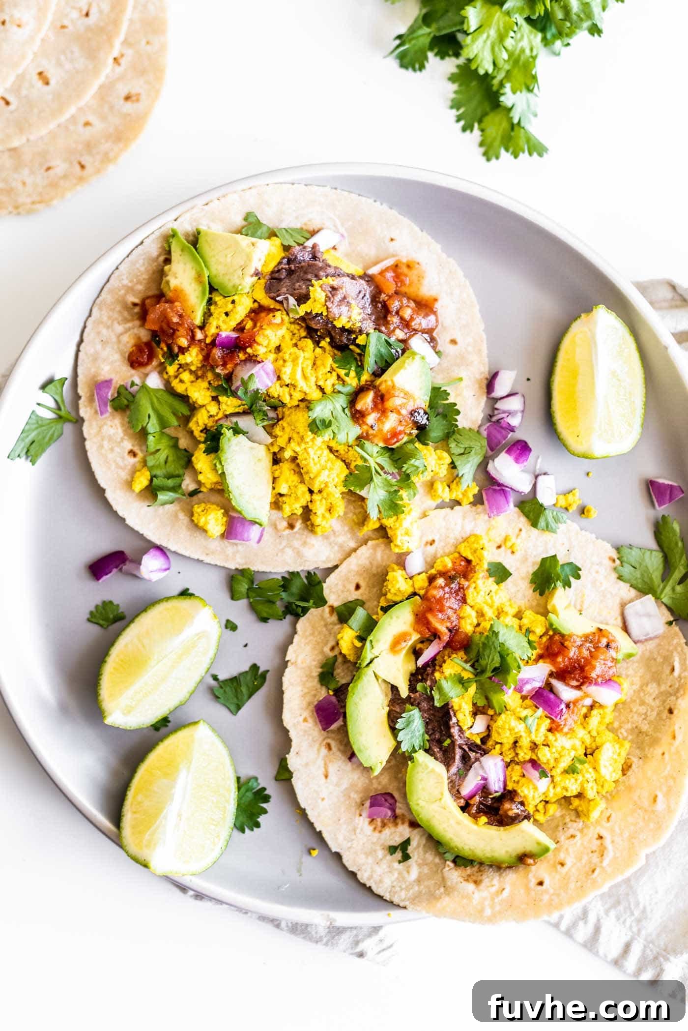 Plant-Powered Breakfast Tacos 2 Two vibrant vegan breakfast tacos arranged on a plate, garnished with fresh lime slices and a sprinkle of cilantro. The tacos are generously filled with fluffy tofu scramble, creamy refried beans, and sliced avocado, promising a flavorful and satisfying start to the day.