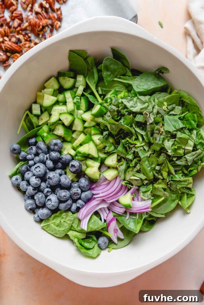 A large bowl filled with the fresh salad ingredients: vibrant spinach, ripe blueberries, thinly sliced red onion, crisp cucumbers, and aromatic basil, ready for dressing.