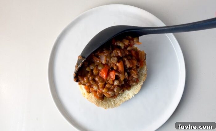 Scooping a lentil sloppy joe mixture over a toasted bun on a plate, ready for serving.