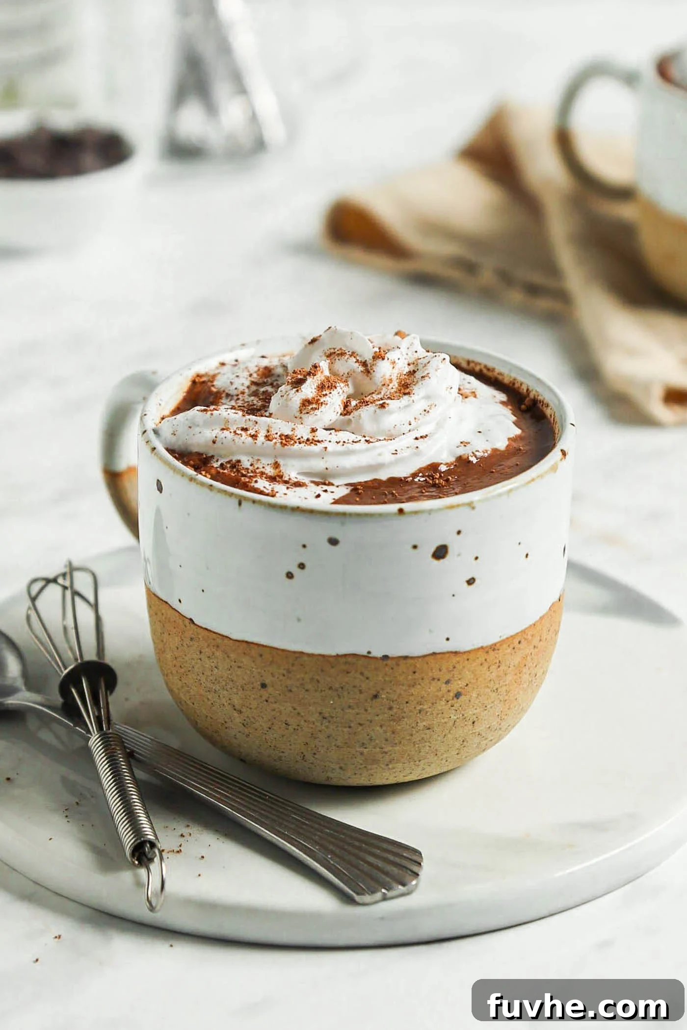 Decadent Plant-Based Hot Chocolate 2 Close up of a mug of hot chocolate topped with whipped cream on a small plate with a spoon and a small whisk.