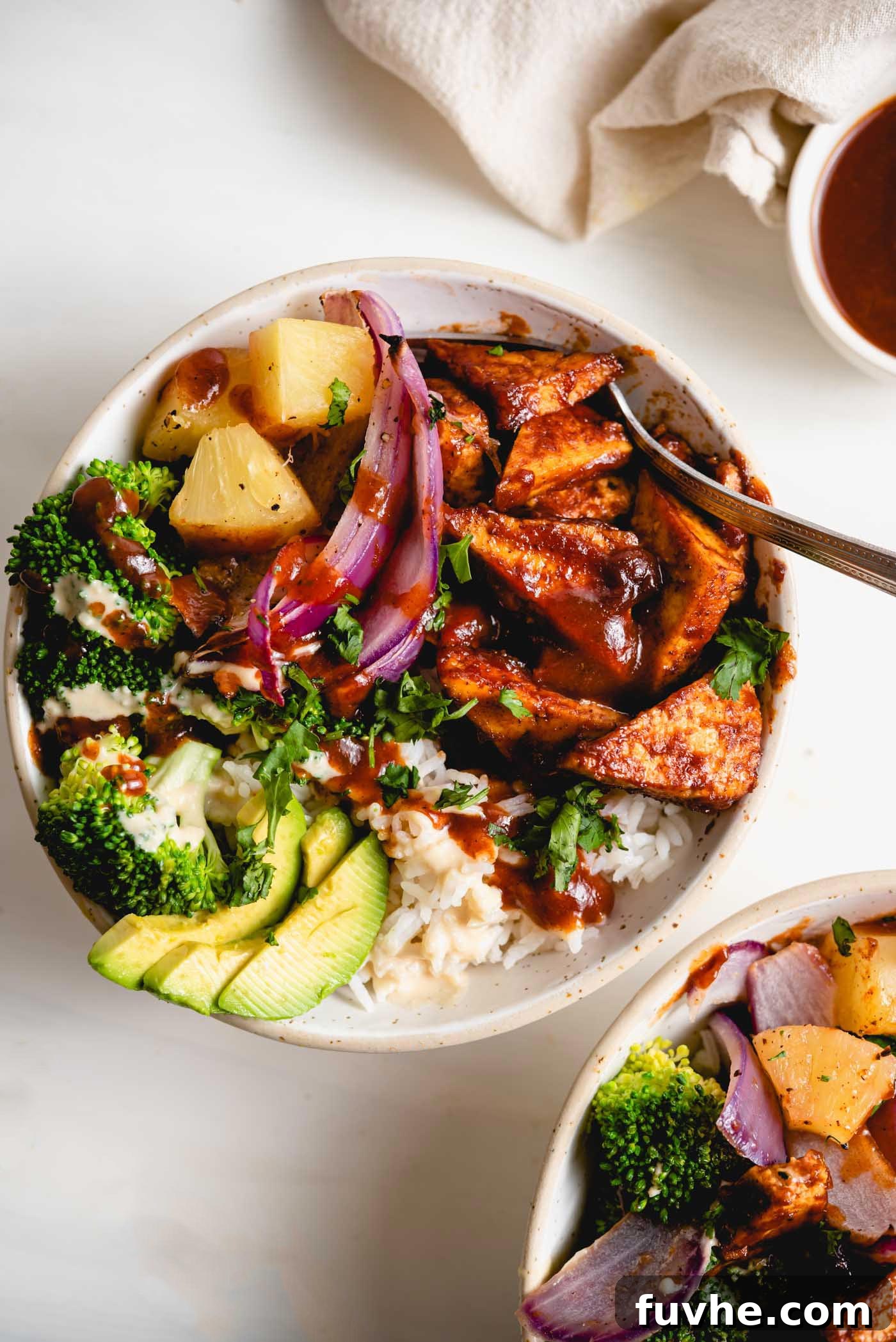A beautifully arranged vegan BBQ tofu bowl with rice, roasted BBQ tofu, fresh avocado, sliced red onion, and juicy pineapple chunks, photographed from an overhead perspective on a light surface.