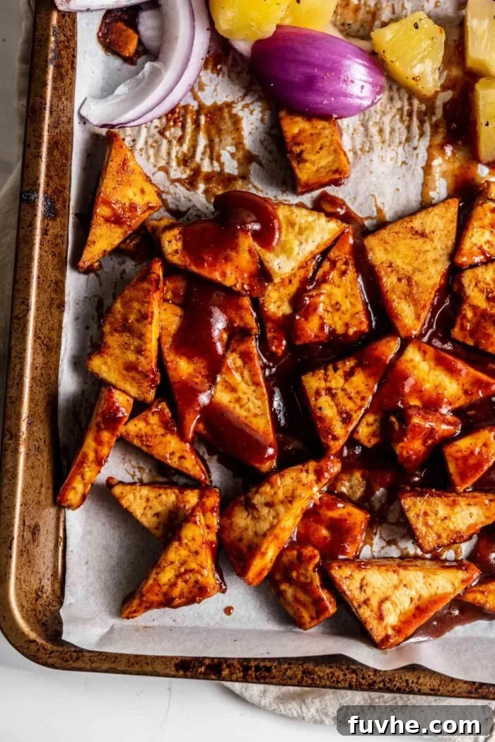 Golden-brown tofu triangles coated in rich BBQ sauce, arranged on one side of a parchment paper-lined baking tray, with sliced red onions occupying the other side, all ready for the final roast.