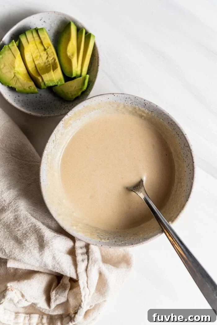 A small, elegant bowl filled with a creamy, light-colored tahini dressing, accompanied by a small spoon, showcasing a perfect topping for any bowl.