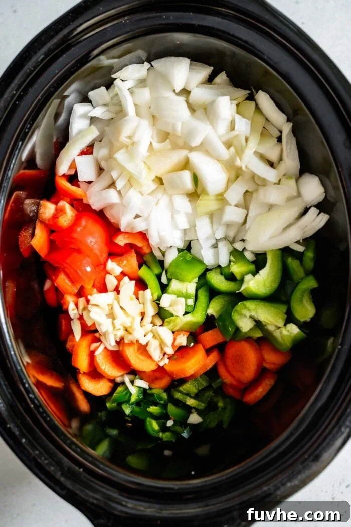 A top-down view of a slow cooker bowl, showcasing a colorful medley of freshly chopped onion, bell peppers, carrot, jalapeño, and garlic, forming the base of the chili.