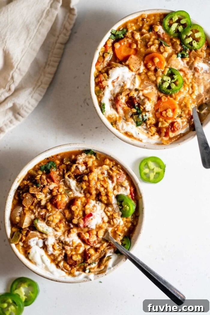 Two inviting bowls of red lentil chili, each beautifully garnished with a swirl of vegan sour cream and a sprinkle of vibrant green sliced jalapeños, presented on a rustic wooden surface.