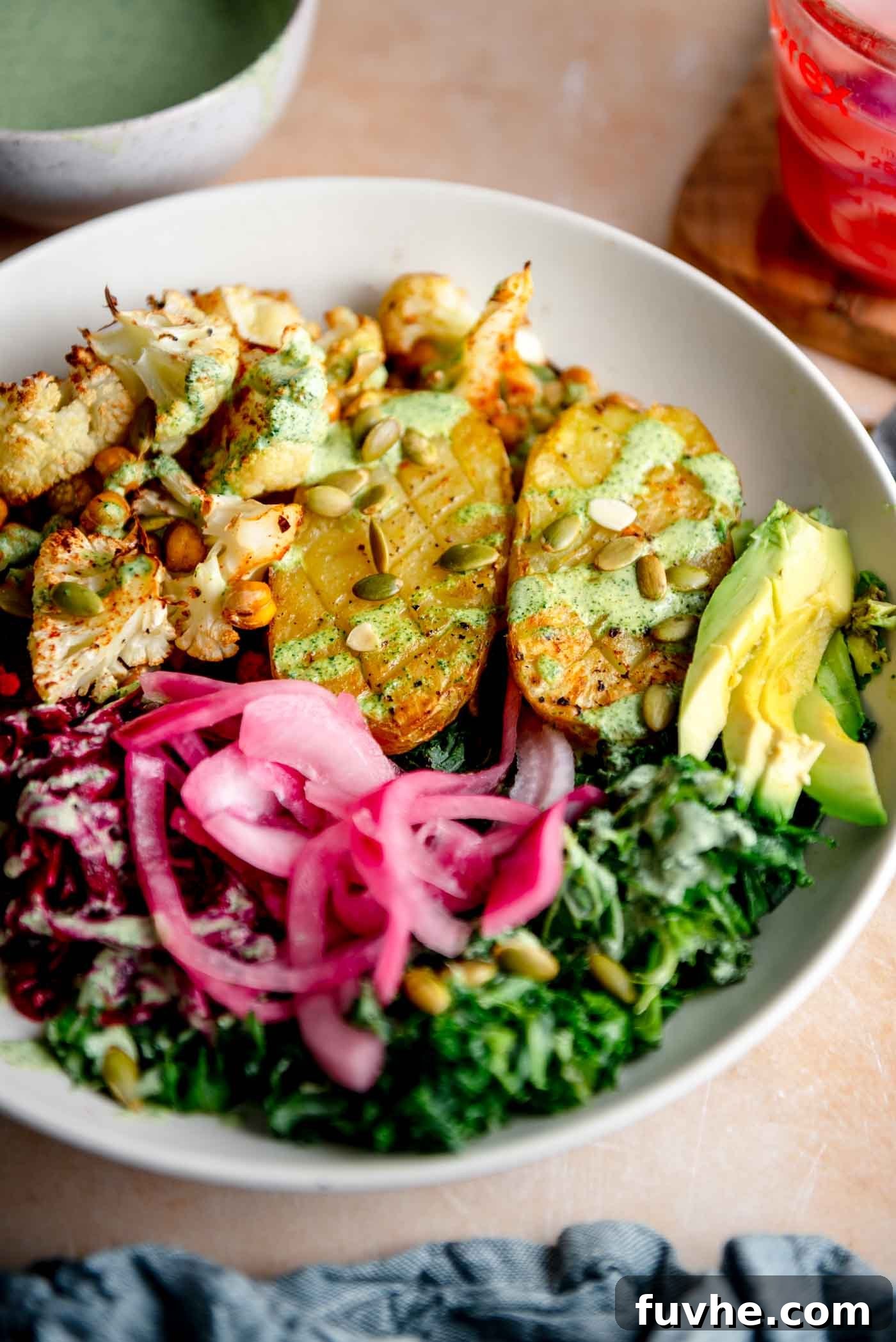 A vibrant bowl featuring a baked potato, crispy roasted cauliflower and chickpeas, fresh kale, pickled red onion, shredded beets, and a creamy green dressing drizzled over top, ready to be enjoyed.