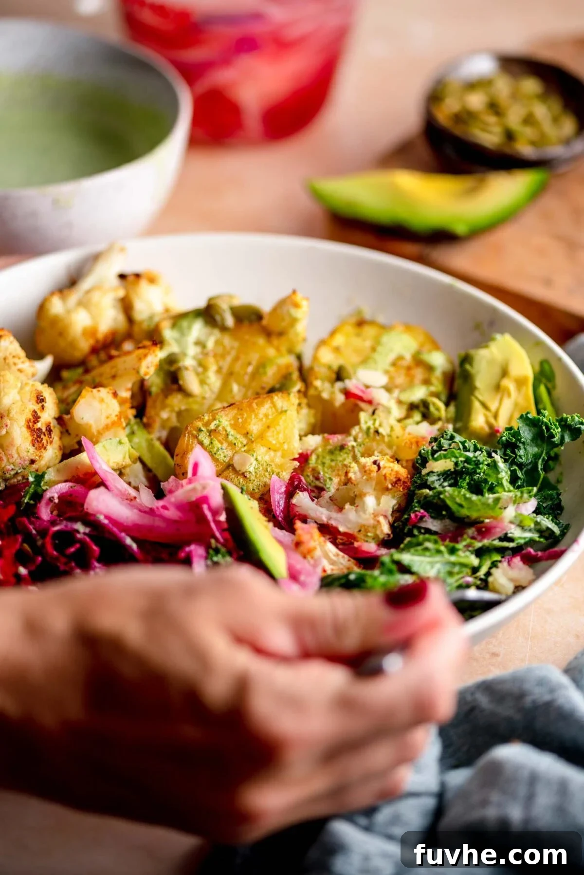 A close-up shot of hands holding a fork, about to enjoy a mouthful of a roasted potato, pickled onion, kale, roasted cauliflower, and chickpeas from a healthy bowl.