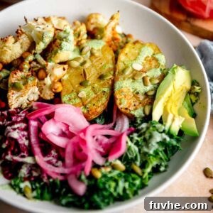 A vibrant bowl featuring a baked potato, crispy roasted cauliflower and chickpeas, fresh kale, pickled red onion, shredded beets, and a creamy green dressing drizzled over top.
