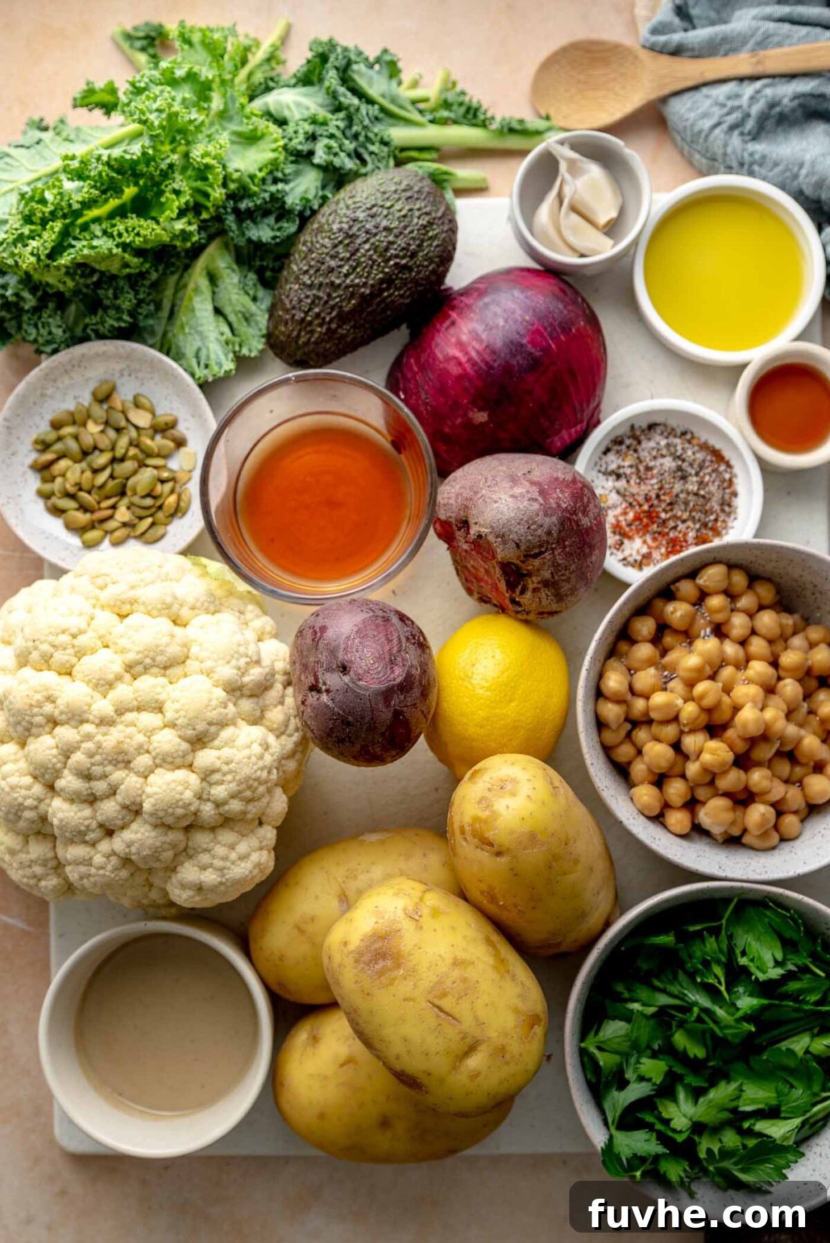 Various fresh ingredients laid out on a wooden cutting board, including cauliflower florets, a bowl of cooked chickpeas, fresh kale leaves, thinly sliced red onion, olive oil, fresh cilantro and parsley, pumpkin seeds, a lemon, and raw beets, all ready for preparing healthy roasted potato bowls.