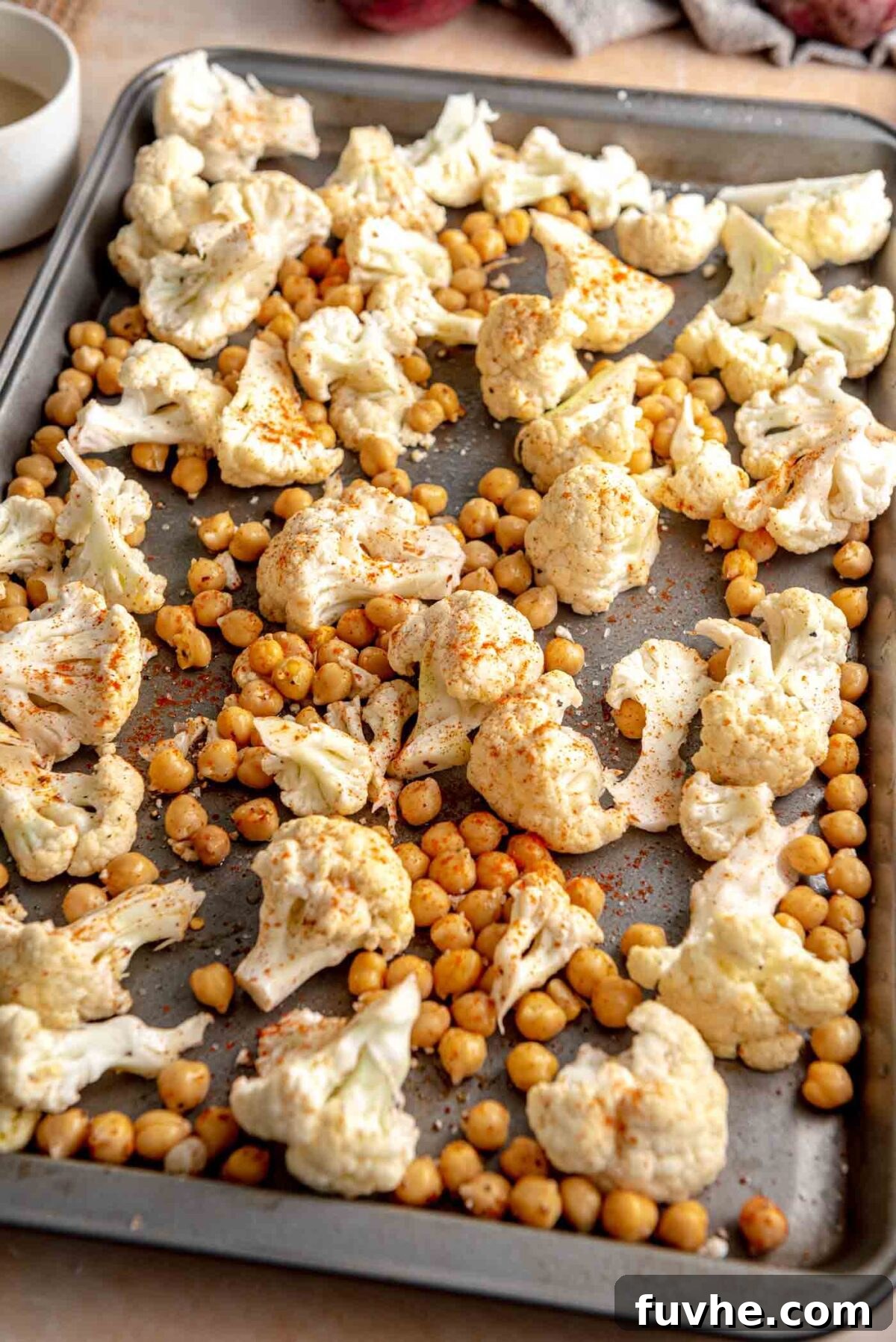 Chopped cauliflower florets and rinsed chickpeas spread on a baking tray, seasoned and ready for roasting.