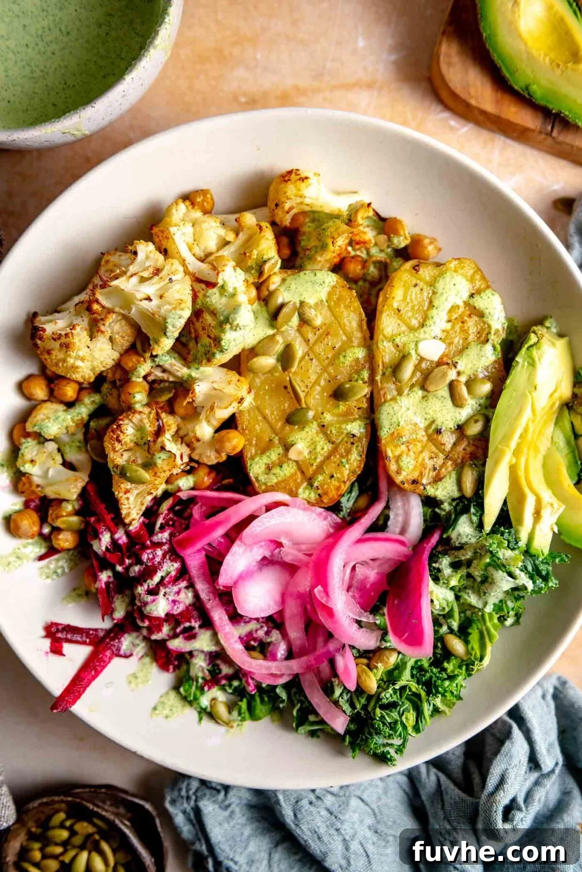 An assembled bowl featuring a baked potato, roasted cauliflower and chickpeas, massaged kale, vibrant pickled red onions, and a generous drizzle of creamy green dressing, ready to be served.