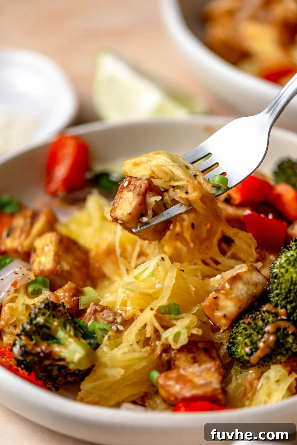 Fork with a piece of tofu and some spaghetti squash on it over a plate of spaghetti squash and roasted vegetables.