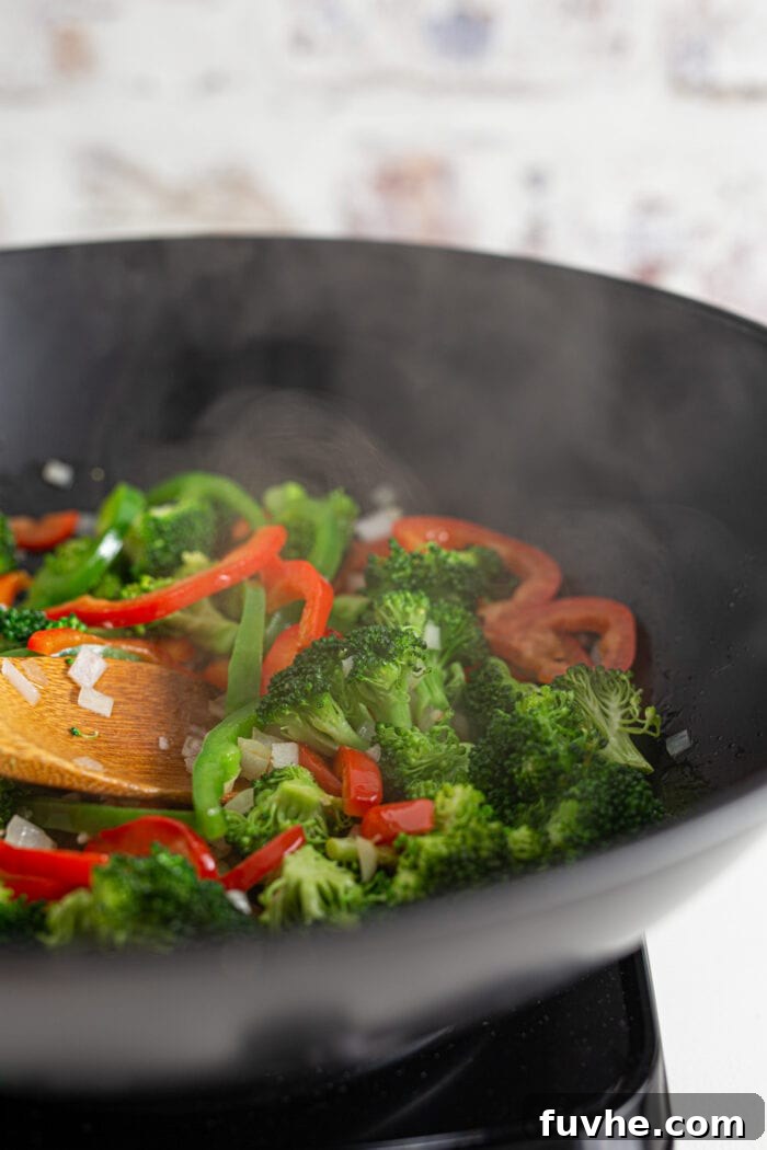 Vibrant broccoli florets and sliced bell peppers are actively cooking in a hot wok, with steam rising from the pan, indicating the sautéing process.