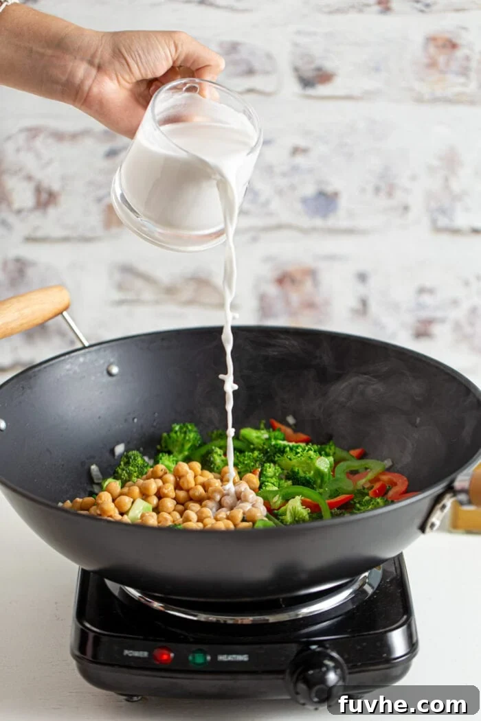 Coconut milk is being poured into a hot wok filled with a mixture of sautéed vegetables and chickpeas, forming the base of the curry sauce.