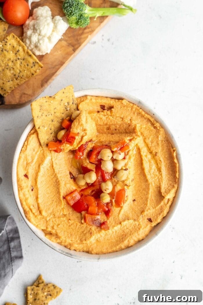 A vibrant bowl of homemade roasted red pepper hummus, garnished with a sprinkle of paprika and fresh parsley, with a crunchy tortilla chip being dipped into its creamy texture.