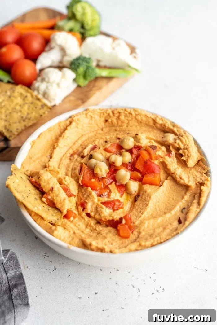 A bowl of vibrant roasted red pepper hummus, accompanied by a selection of fresh, crisp chopped vegetables like carrots, cucumbers, and bell peppers, ready for dipping.