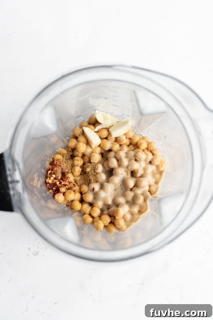 Raw chickpeas, tahini, whole garlic cloves, and a lemon half are placed together in a high-speed blender, ready to be processed for homemade hummus.