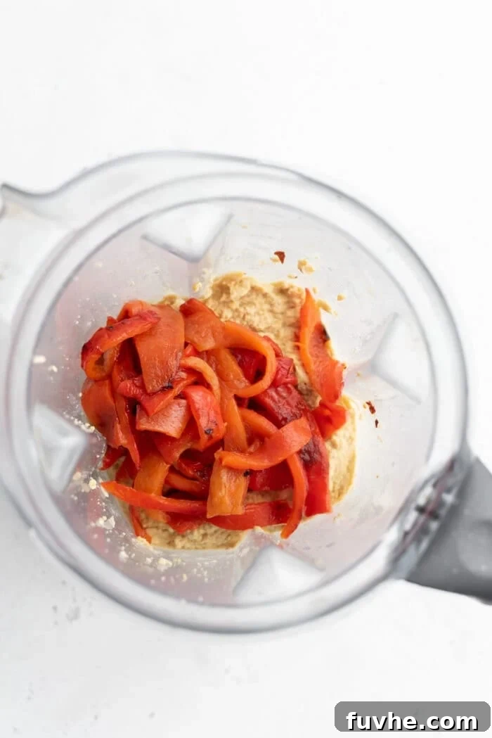 Freshly roasted red peppers, with their charred skins removed, are added to a blender alongside the creamy hummus base, poised for blending into a vibrant dip.