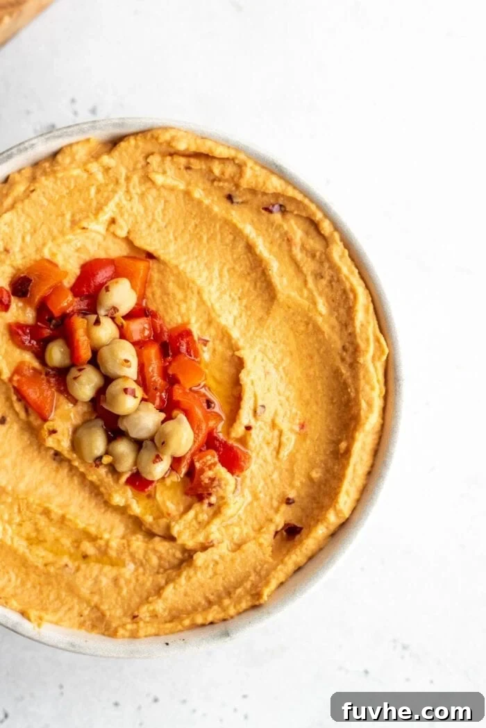 An overhead close-up view of a beautifully garnished bowl of roasted red pepper hummus, topped with whole chickpeas, finely diced roasted red pepper pieces, and a sprinkle of vibrant red pepper flakes.