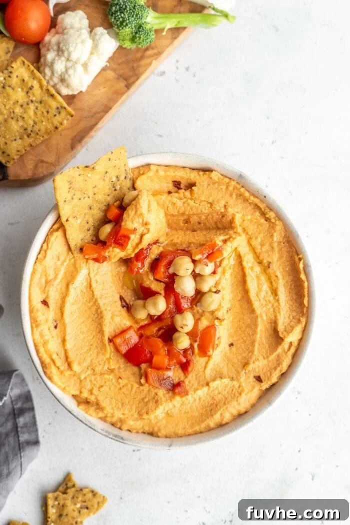 A bowl of vibrant homemade roasted red pepper hummus with a tortilla chip partially dipped into its smooth texture, highlighting its creamy consistency.