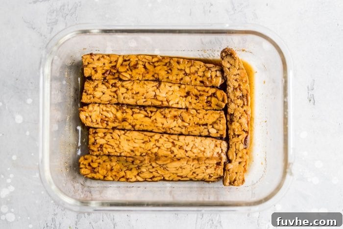 Five strips of thinly sliced tempeh marinating in a clear glass dish, absorbing the rich, dark marinade, ready for transformation into delicious tempeh bacon.