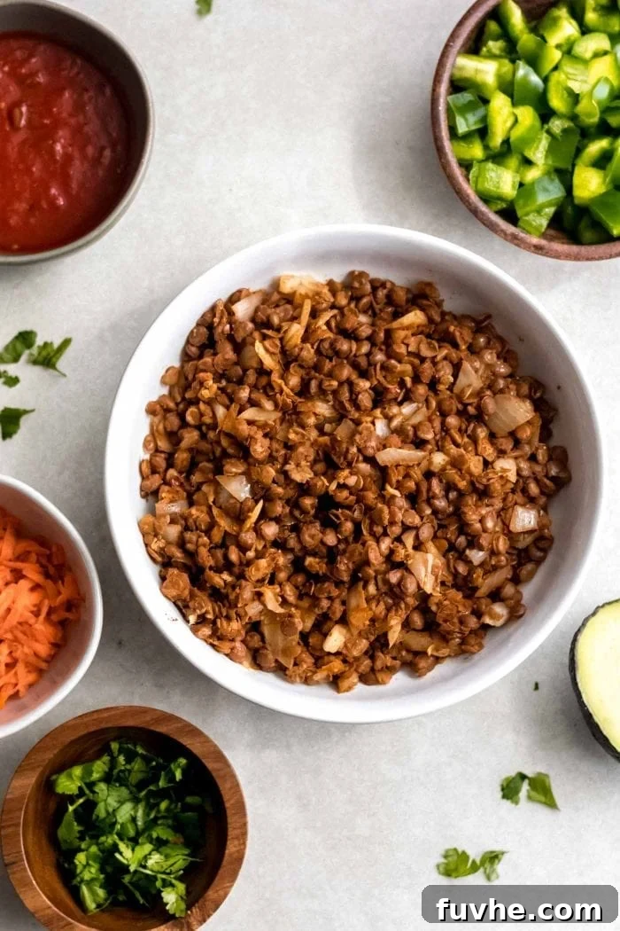 A beautiful spread of vegan lentil taco components: a white bowl overflowing with lentil filling, surrounded by bowls of fresh cilantro, crunchy carrots, creamy avocado slices, and zesty salsa.