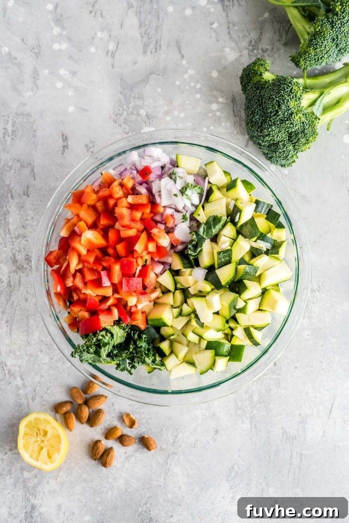 Vibrant Chickpea Garden Salad 5 Large mixing bowl filled with a colorful medley of zucchini, bell pepper, kale, and red onion. Beside the bowl are two pieces of broccoli and some chopped almonds.