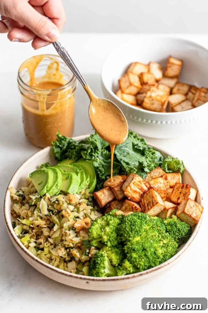 Wholesome Vegan Carb-Conscious Bowls 12 A hand drizzling a spoonful of creamy satay sauce over a fully assembled vegan dinner bowl filled with tofu, avocado, and various vegetables.