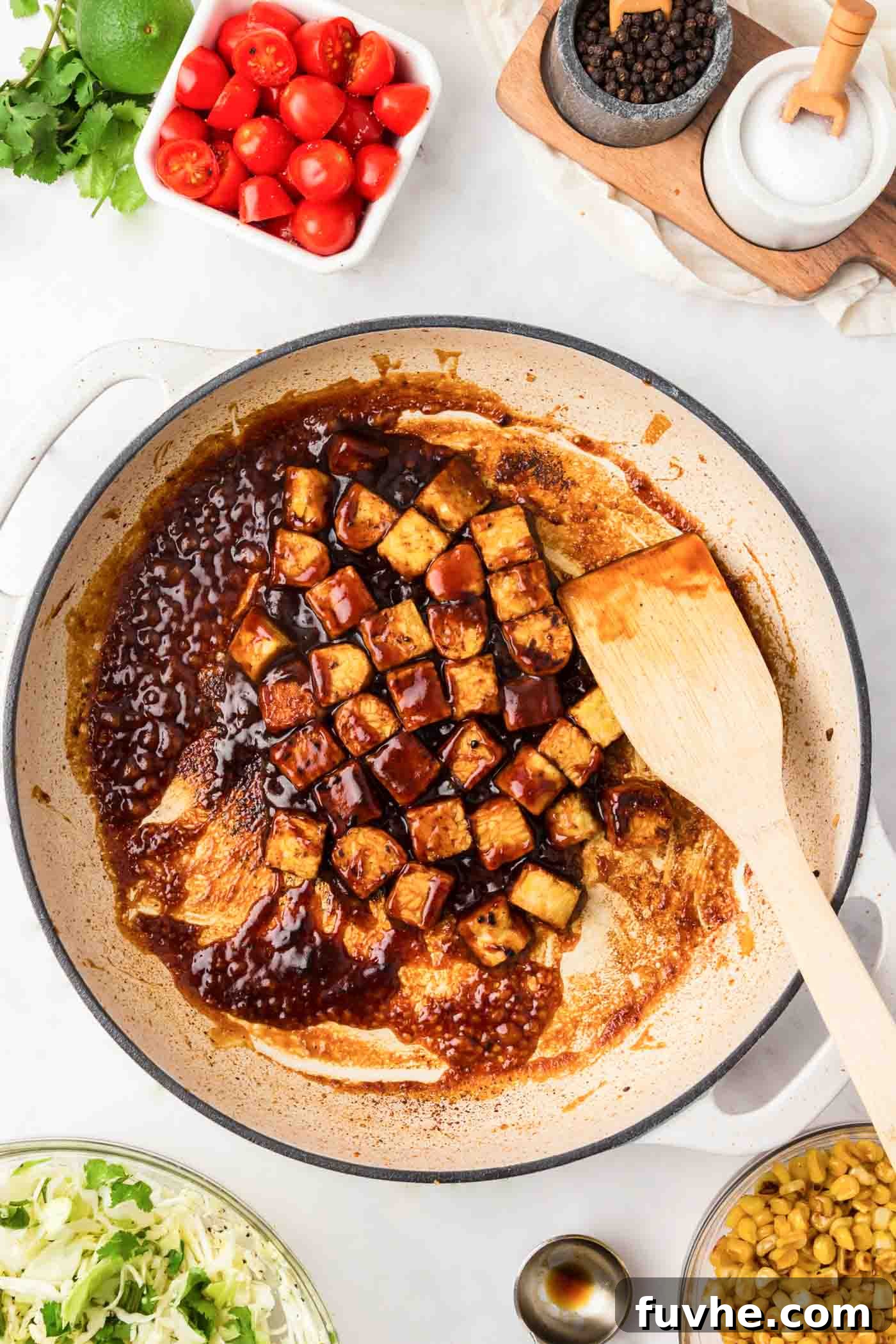 Cubes of tempeh coated in a rich BBQ sauce, simmering in a pan with a wooden spoon.