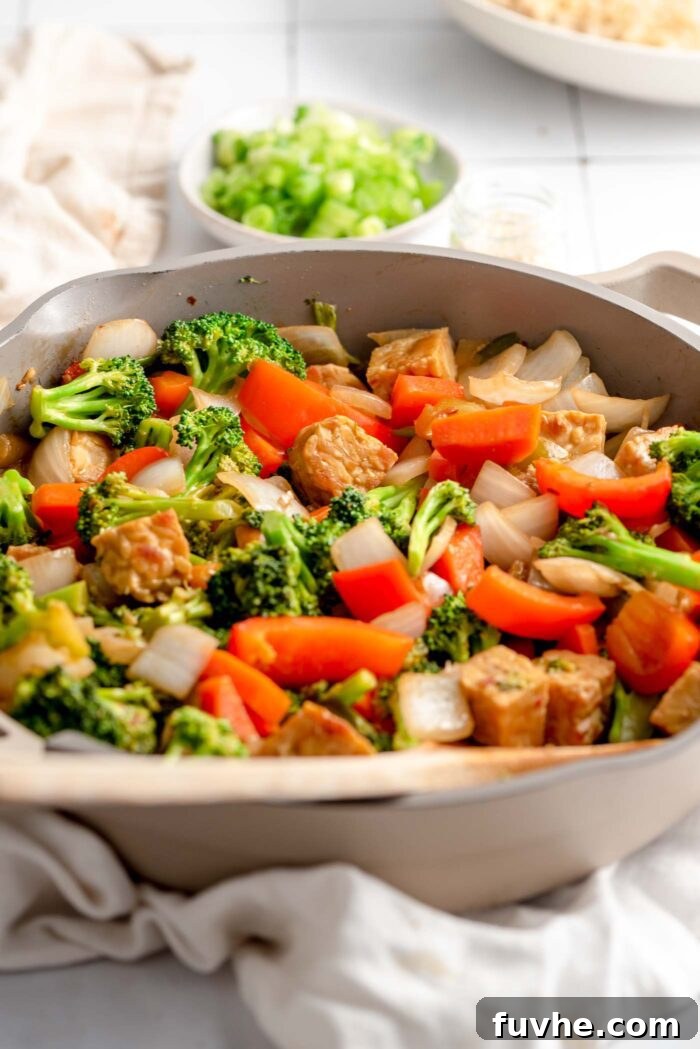 Golden Tempeh Wok Medley 7 A finished vegetarian stir fry with crispy tempeh, broccoli, red bell pepper, and onion, richly coated in sauce, being stirred in a skillet. A small dish of chopped green onions sits in the background, ready for garnish.