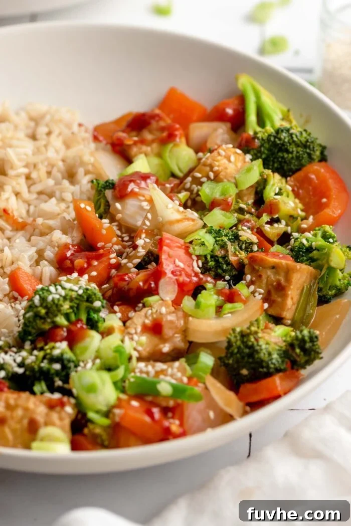 Golden Tempeh Wok Medley 8 An inviting angled view of a bowl filled with vegan tempeh and vegetable stir fry, served over brown rice. The dish is beautifully topped with toasted sesame seeds and vibrant chopped scallions, ready to be enjoyed.