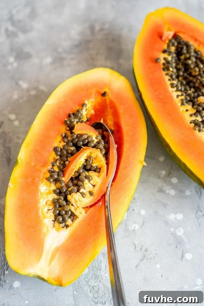 A spoon delicately scooping out the black seeds from the center of a freshly cut papaya half, illustrating the preparation process.