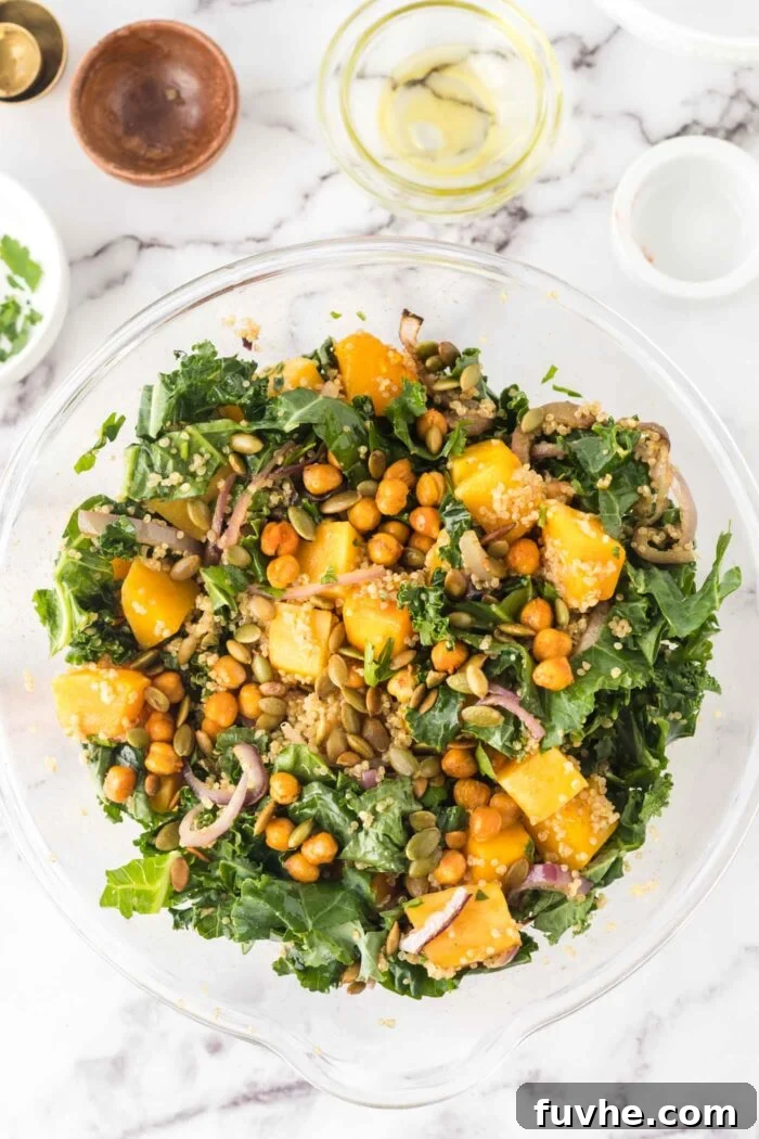 A partially assembled kale salad featuring quinoa, red onion, roasted butternut squash, and chickpeas, being tossed in a large mixing bowl.