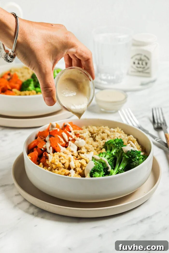 Hand pouring tahini sauce from a small jar over a bowl with lentils, quinoa, roasted vegetables and broccoli, completing the assembly.