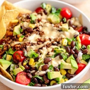 A bowl of vegan taco salad with a creamy dressing drizzled over vegan taco crumbles, lettuce, tomato, avocado, black beans, red onion and corn. There are some tortilla chips on the side.