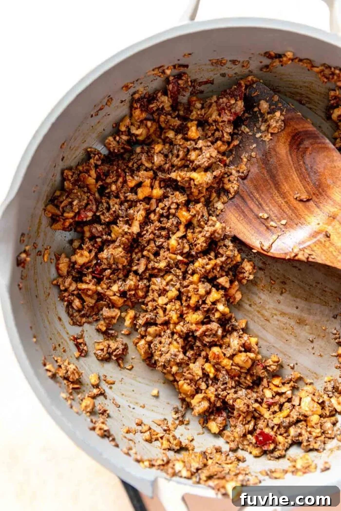 Savory mushroom and walnut crumbles cooking in a skillet with spices, stirred with a wooden spoon until browned and tender.