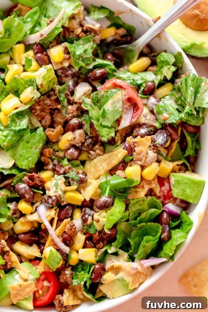 A large, vibrant vegan taco salad bowl, overflowing with savory walnut-mushroom crumbles, crisp lettuce, sweet corn, thinly sliced red onion, juicy tomatoes, creamy avocado, and a generous drizzle of creamy dressing, served with crunchy tortilla chips.