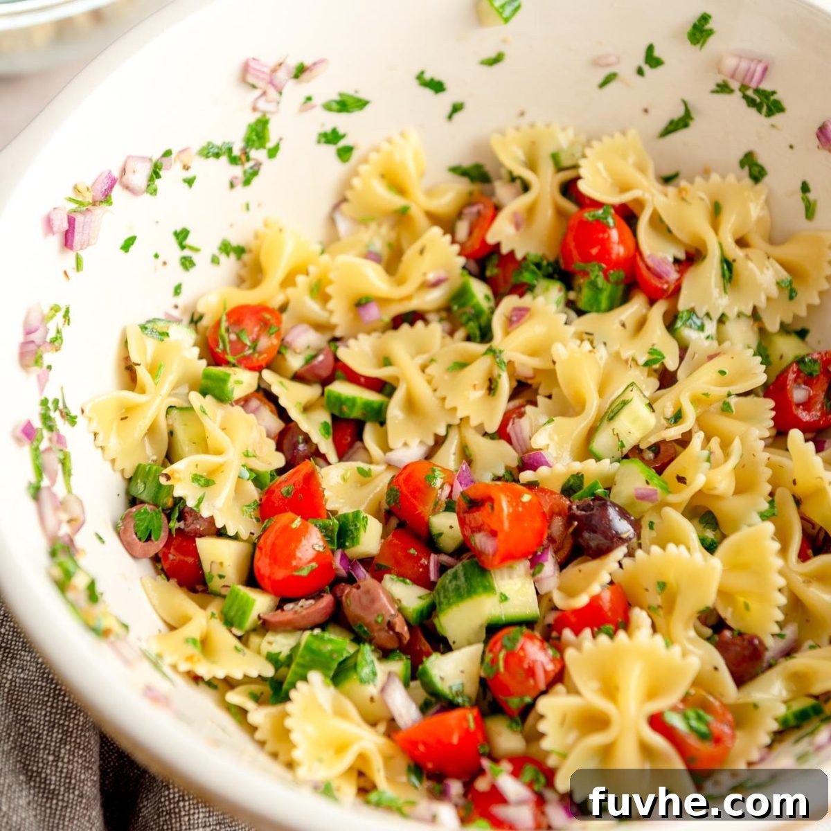 Plant-Based Greek Pasta Salad 7 Greek pasta salad made with bow-shaped pasta, cucumber, tomato, olives and red onion in a large mixing bowl.